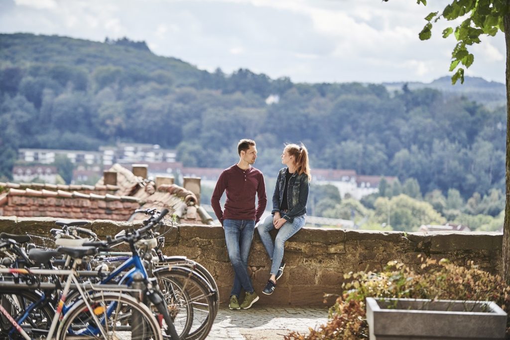 230923 HT LAHNTAL MARBURG 1700 FINAL WEB Large Foto von einem Mann und einer Frau vor einer Mauer. Im Hintergrund ist ein Panoramablick über Marburg zu sehen und im Vordergrund stehen Räder.